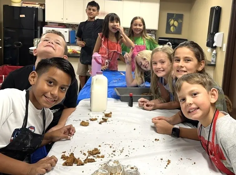 Smiling group of children gathered around a table while baking and decorating treats in a kitchen classroom.